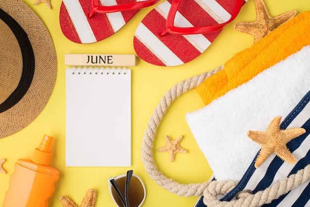 Top view photo of copybook with inscription june starfishes beach bag with orange and white towels sunscreen bottle cap sunglasses and flipflops on isolated yellow background with blank space