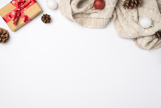 Top view photo of craft paper giftbox with red ribbon bow cozy knitted pullover pine cones red and white christmas tree balls on isolated white background with copyspace