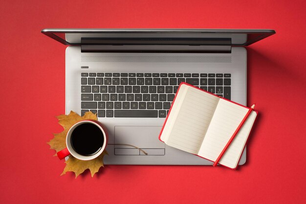 Top view photo of cup of coffee on autumn orange maple leaf open red planner and pencil on open grey laptop on isolated red background