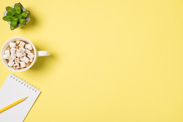 Top view photo of cup of drink with marshmallow plant and\
yellow pencil on small spiral copybook on isolated light yellow\
background with copyspace