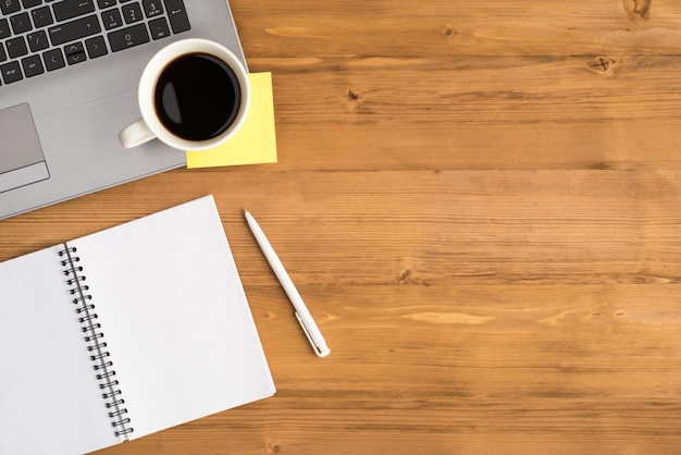 Top view photo of cup of drink yellow sticker note paper on laptop pen and open spiral notepad on isolated wooden table background with blank space