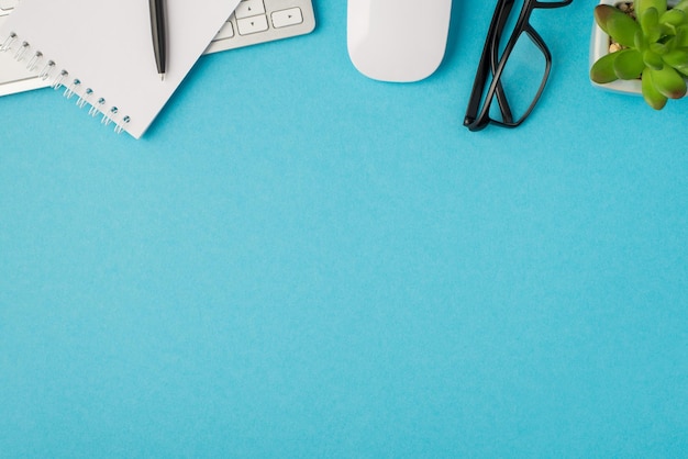 Top view photo of desktop with keyboard pen on notepad glasses mouse and plant on isolated blue background with empty space