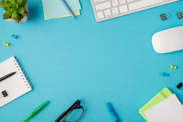 Top view photo of desktop with keyboard pens clips organizes pencil on stickers pen and clip on notebook glasses mouse and plant on isolated blue background with copyspace