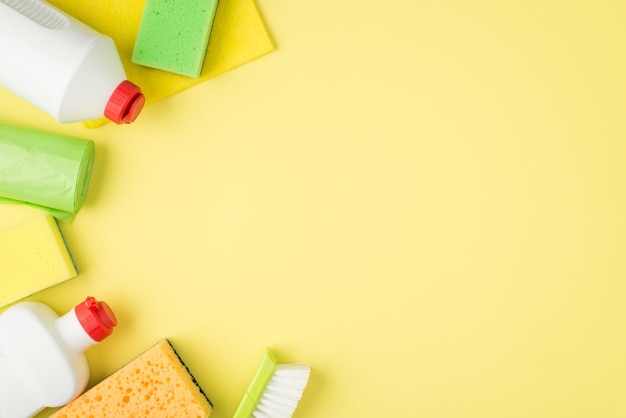 Top view photo of detergent plastic bottles yellow and green sponges viscose rag brush and garbage bags on isolated yellow background with copyspace on the right