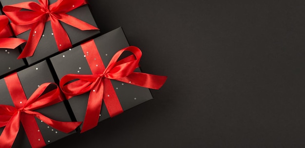 Top view photo of gift boxes in black packaging with sequins and red ribbon bow on isolated black background with empty space