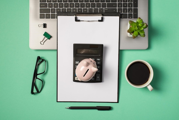 Top view photo of green and black binder clips flowerpot on grey laptop glasses cup of coffee pen black clipboard folder with paper sheet and piggy bank on calculator on isolated turquoise background