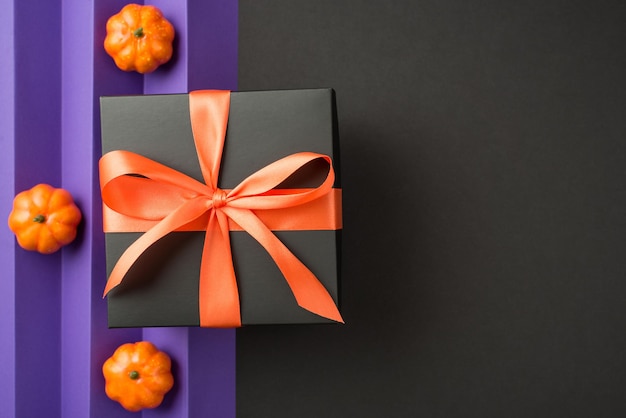 Вид сверху фото композиции на Хэллоуин, черная подарочная коробка с оранжевым бантом из ленты и маленькими тыквами на изолированной спине и фиолетовым листом с вертикальными складками фона с копирайтом