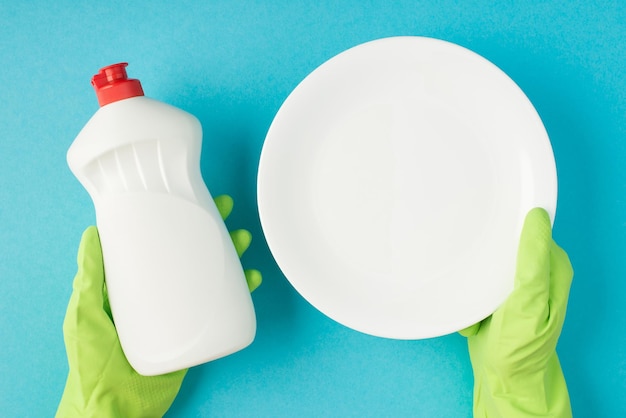 Top view photo of hands in green rubber gloves holding white clean plate and detergent gel bottle without label on isolated pastel blue background with copyspace