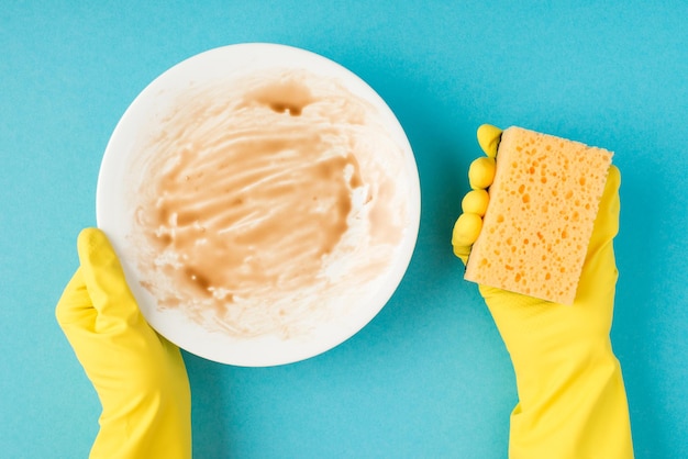 Top view photo of hands in yellow rubber gloves holding dirty plate and scouring pad on isolated pastel blue background