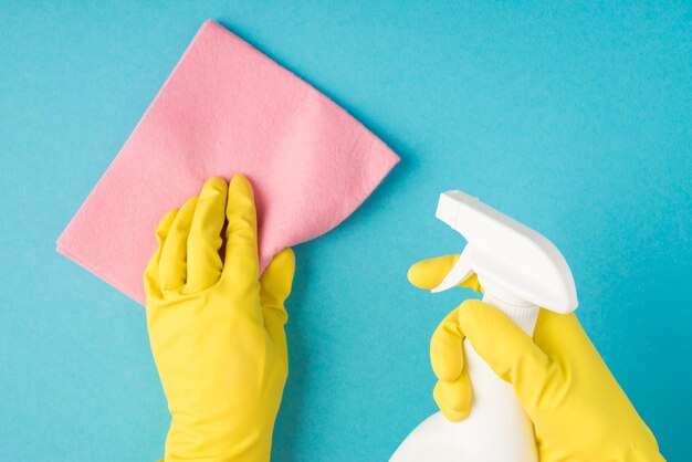 Top view photo of hands in yellow rubber gloves holding pink viscose rag and white spray bottle on isolated pastel blue background