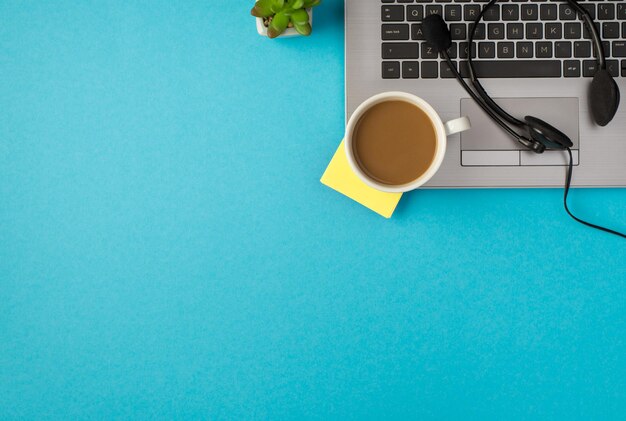 Top view photo of headset with microphone cup of tea sticker note on laptop and plant on isolated pastel blue background with blank space