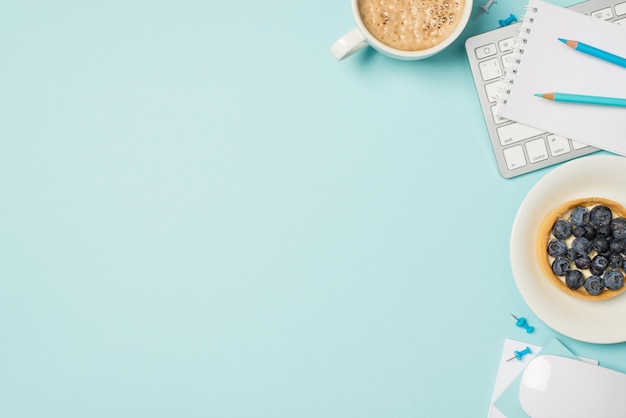 Top view photo of keyboard mouse planner blue pencils pins plate with blueberry cake and cup of frothy latte on isolated pastel blue background with empty space