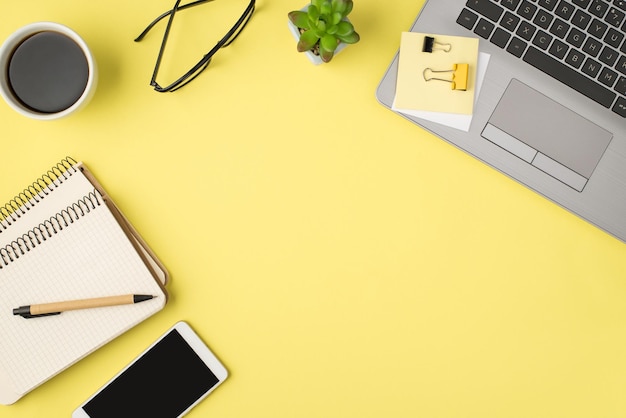 Top view photo of laptop glasses flowerpot pen binder clips\
sticker note paper cup of coffee mobile phone and two spiral\
copybooks on isolated pastel yellow background with empty\
space