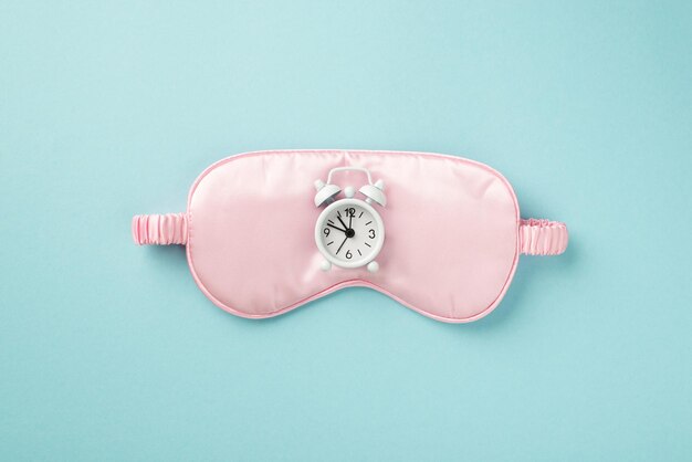 Top view photo of the little white alarm and big pink silk sleep mask in the middle of the isolated pastel blue background copyspace