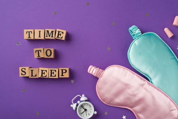 top view photo of one white clock and two silk blindfolds pink and light blue one with scattered confetti in shape of stars wooden cubes with letters and two earplugs on the violet background