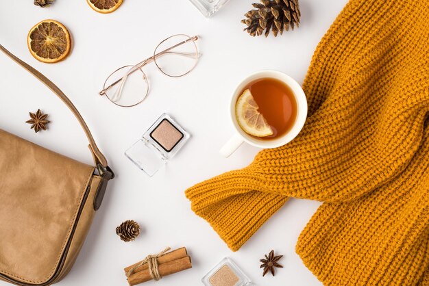 Top view photo of orange sweater cup of tea with lemon eyeshadows cosmetics stylish glasses pine cones anise dried lemon slices cinnamon sticks and leather handbag on isolated white background