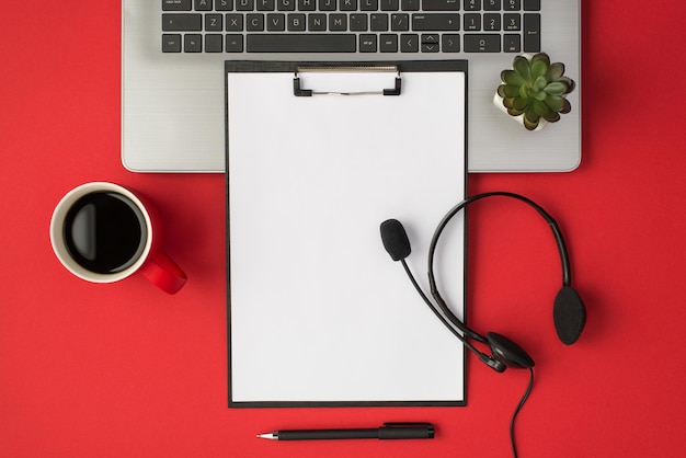 Top view photo of plant on laptop cup of coffee pen black clipboard folder with white paper sheet and headset with microphone on isolated red background with copyspace