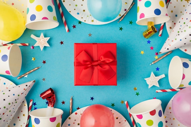 Top view photo of red gift box with ribbon bow in the middle of birthday table composition and party accessories on isolated blue background