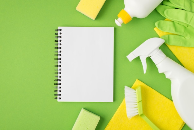 Photo top view photo of reminder in the middle detergent spray and gel bottles sponges brush yellow napkins green rubber gloves on isolated green background with copyspace