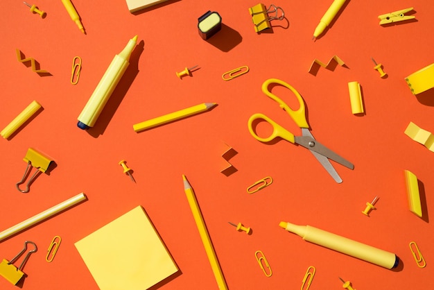散在する黄色の文房具学校の付属品はさみ鉛筆マーカーバインダークリップ画鋲と孤立した鮮やかなオレンジ色の背景に付箋紙の上面写真