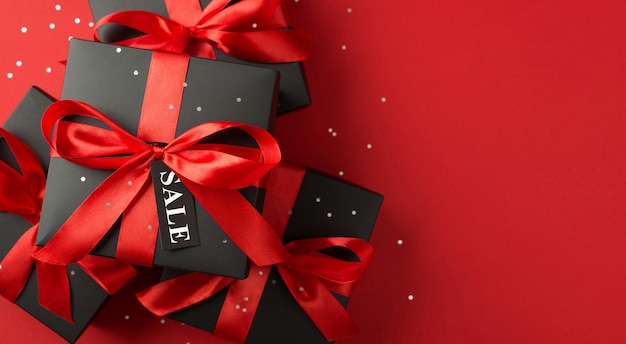 Top view photo of sequins stack of black gift boxes with red ribbon bow and tag on isolated red background with text sale on pricetag