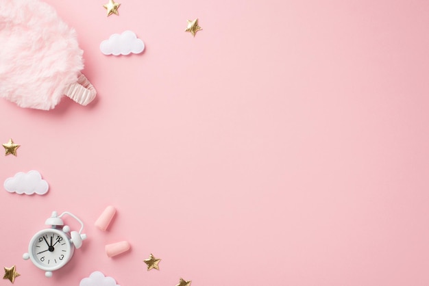 Top view photo of small white alarm clock pink fluffy sleeping mask earplugs golden stars and clouds on isolated pastel pink background with empty space