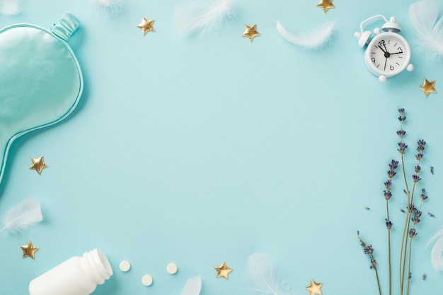 Top view photo of sprig of lavender feathers small white alarm clock blue silk sleeping mask open bottle with pills and golden stars on isolated pastel blue background with copyspace in the middle