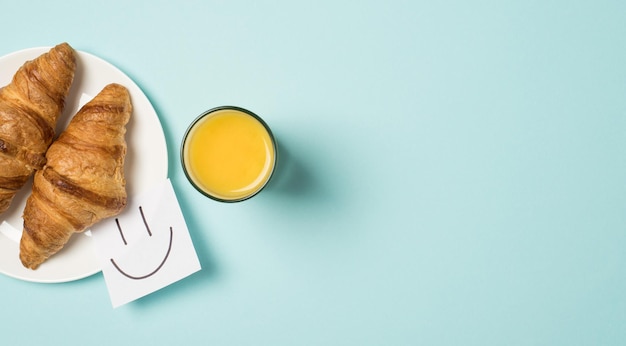 Top view photo of sticker note paper with drawn smiling face glass of juice and plate with two fresh croissants on isolated light blue background with copyspace