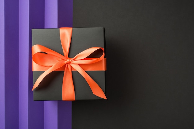 Top view photo of stylish black giftbox with orange ribbon bow on isolated back and violet sheet with vertical folds background with empty space