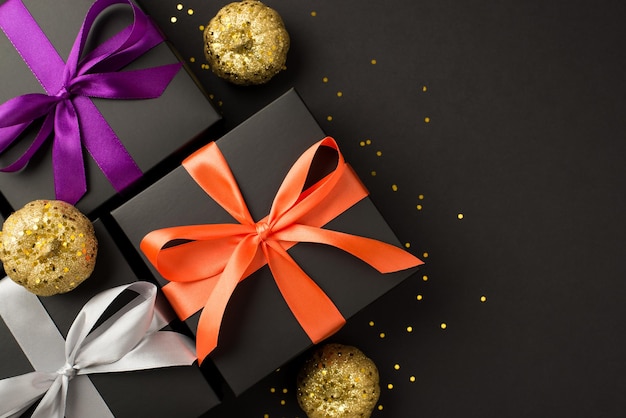 Top view photo of three black gift boxes with violet orange and white ribbon bows golden confetti and shiny golden pumpkins on isolated black background with blank space