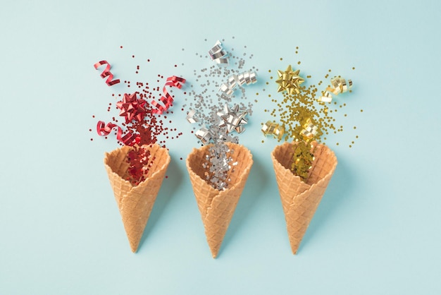 Top view photo of three ice cream cones with christmas decorations explosion of red silver and gold sequins serpentine and stars on isolated pastel blue background