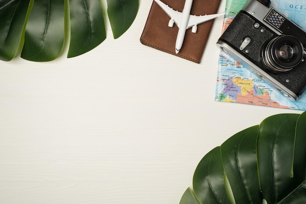 Top view photo of two green palm leaves plane model on passport cover camera and map on wooden white background with copyspace