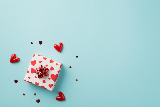 top view photo of valentine's day decor giftbox in white wrapping paper with heart pattern and red star bow surrounded by small hearts and confetti on isolated pastel blue background with blank space