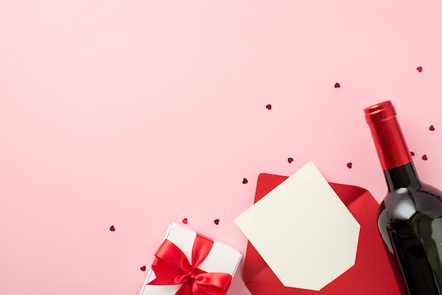 バレンタインデーの装飾ハート型の紙吹雪白いギフト ボックスと赤い弓赤い封筒と紙のシートと空のスペースで分離されたパステル ピンクの背景にワインのボトルの上から見た写真