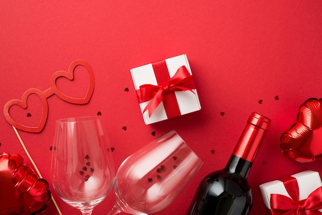 Top view photo of valentine\'s day decorations gift box heart\
shaped paper glasses balloons two wineglasses wine bottle and red\
confetti on isolated red background with copyspace