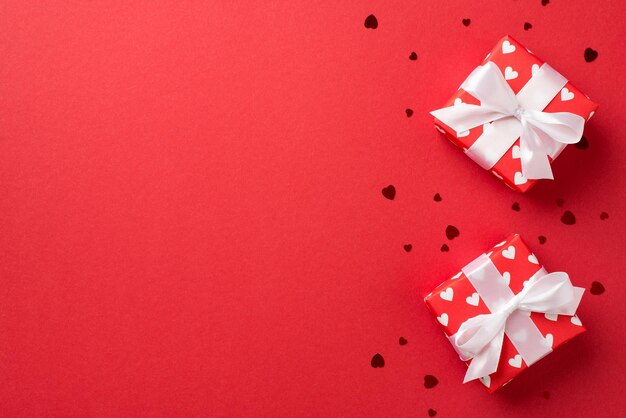 Top view photo of valentine's day decorations gift boxes in red wrapping paper with pattern of hearts white ribbon bows and confetti on isolated red background with copyspace