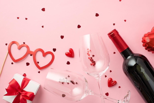 Top view photo of valentine's day decorations giftbox paper heart shaped glasses balloon small hearts two wineglasses with confetti and wine bottle on isolated pastel pink background