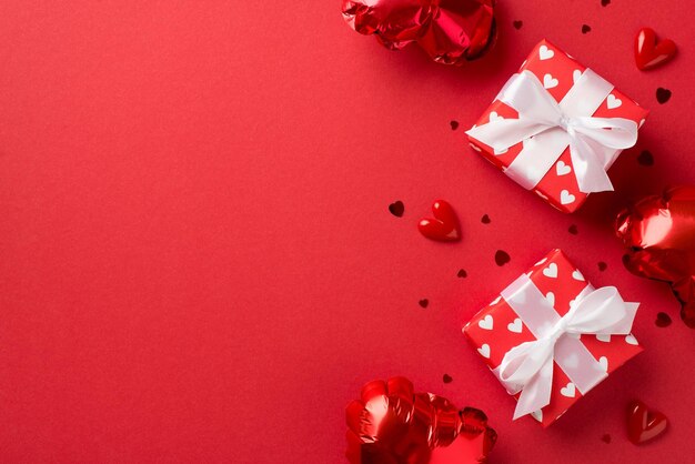 Top view photo of valentine\'s day decorations white gift boxes\
with red ribbon bows heart shaped balloons and confetti on isolated\
red background with empty space