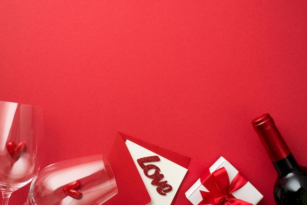 Top view photo of valentine's day decorations white giftbox
with bow small hearts in two wineglasses envelope card with
inscription love and wine bottle on isolated red background with
empty space