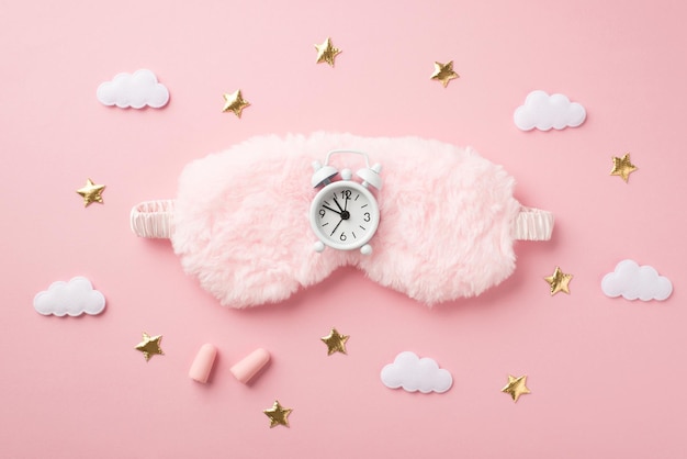 Top view photo of white alarm clock on pink fluffy sleeping mask earplugs clouds and golden stars on isolated pastel pink background