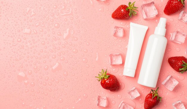 Top view photo of white cream tube and spray bottle without label strawberries ice cubes and drops on the right on isolated pastel pink background with blank space on the left