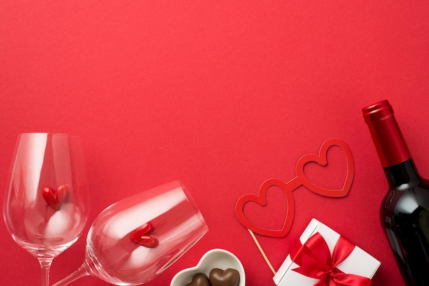 Top view photo of white giftbox with red bow small hearts in two wineglasses heart shaped saucer with candies paper glasses and wine bottle on isolated red background with empty space