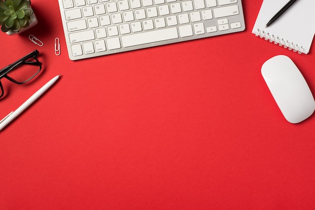 Top view photo of white keyboard mouse flowerpot glasses clips pens and reminder on isolated vivid red background with copyspace