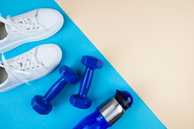 Top view photo of white sneakers water bottle and blue dumbbells on blue beige background with copy space