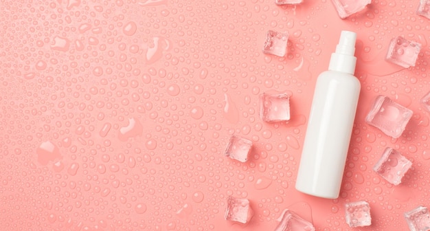 Top view photo of white spray bottle without label ice cubes on the right and drops on isolated pastel pink background with copyspace on the left