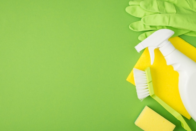 Photo top view photo of white spray detergent bottle sponge brush yellow napkin and green rubber gloves on isolated green background with copyspace on the left