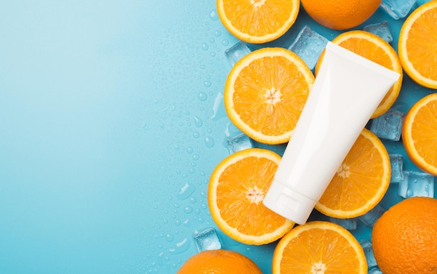 Top view photo of white tube without label on ice cubes water drops halves and whole oranges on isolated pastel blue background with blank space on the left