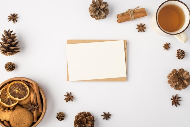 Top view photo of winter composition envelope white card pine cones anise cup of tea wooden bowl with cookies cinnamon sticks and dried lemon slices on isolated white background with blank space