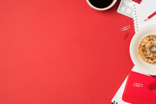 Top view photo of workplace keyboard clips red pencil red copybooks cup of coffee and plate with decorated glazed donut on isolated red background with copyspace