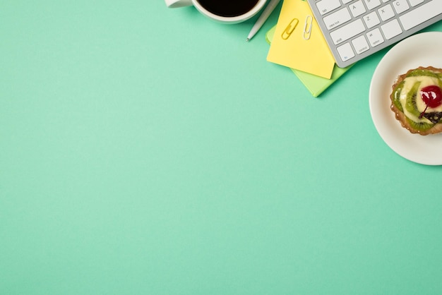 Top view photo of workplace keyboard sticky note paper clips cup of drink and plate decorated cupcake on isolated turquoise background with blank space